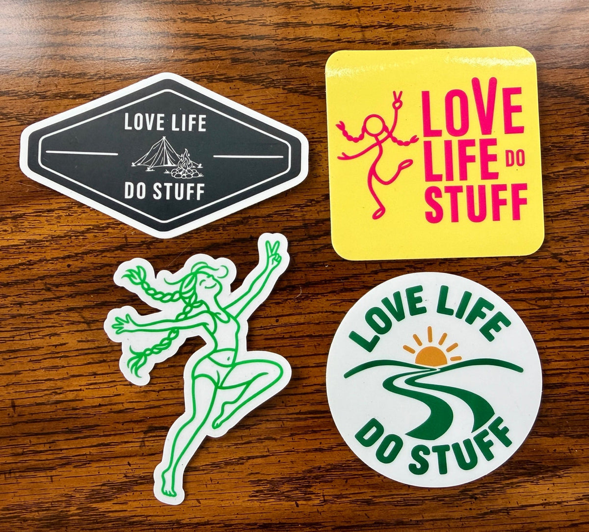 Four Love Life Do Stuff stickers on a wooden table: black camping badge, pink and yellow square, green dancing girl, and green sunrise circle.