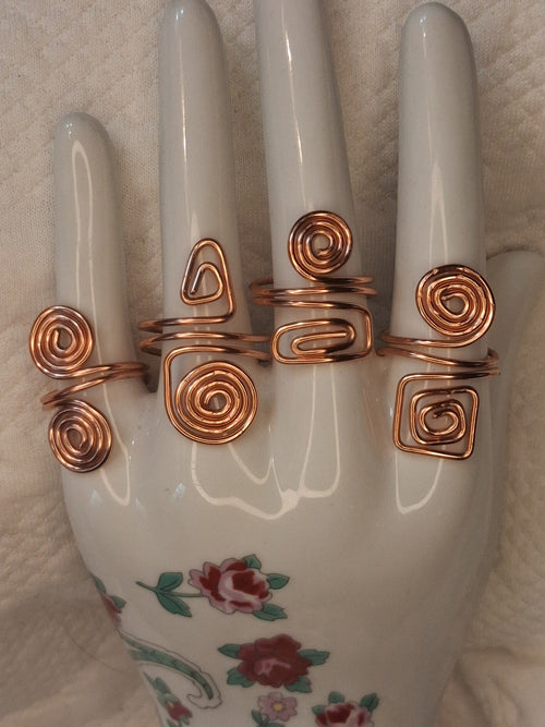 Four different handmade adjustable copper wire rings with spiral, triangular, and rectangular designs on a white jewelry display. Rings sold individually.