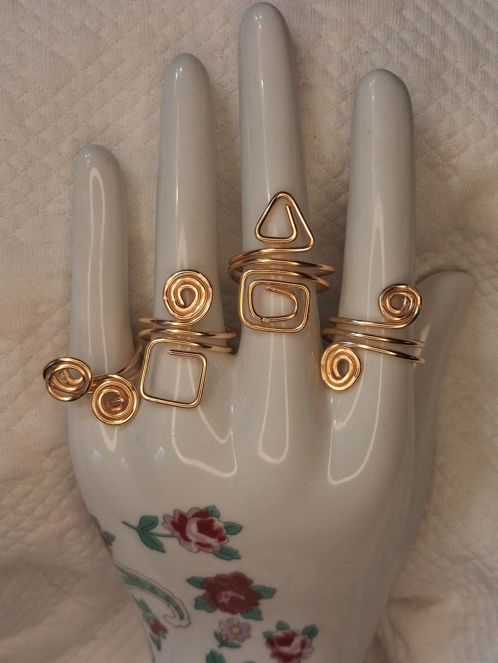 Four handmade adjustable gold wire rings with spiral and geometric designs on a white floral jewelry hand display.