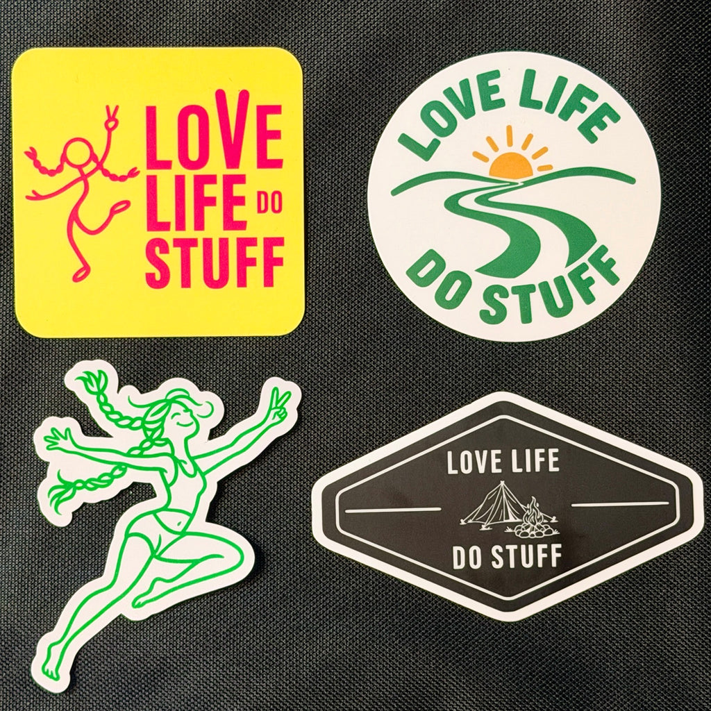 All four Love Life Do Stuff stickers on black fabric: pink and yellow stick figure, round green Wanderlust Badge, green Dancing BeaGrl, and black Vintage Camp Badge.