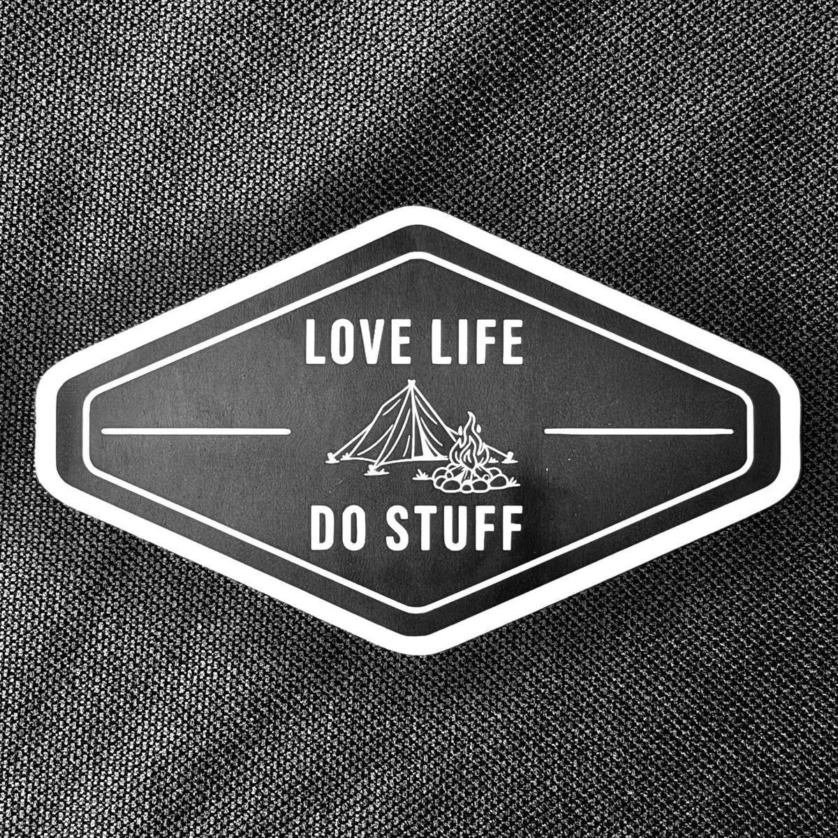 Vintage Camp Badge vinyl sticker on black fabric, with a white tent, campfire, border, and Love Life Do Stuff lettering on a black diamond shape.