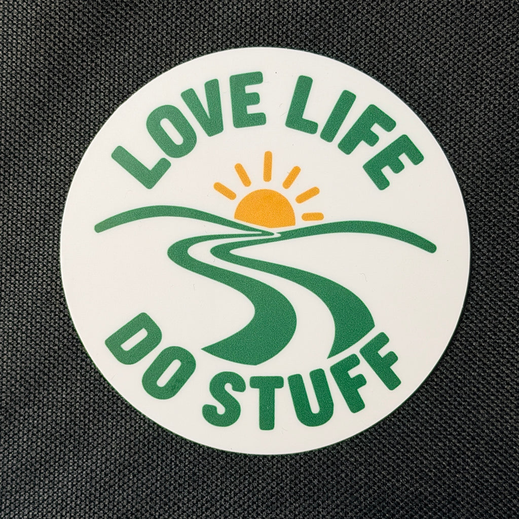 Round Wanderlust Badge vinyl sticker on black fabric, featuring green Love Life Do Stuff lettering, a winding green path, and a golden sun on a white background.