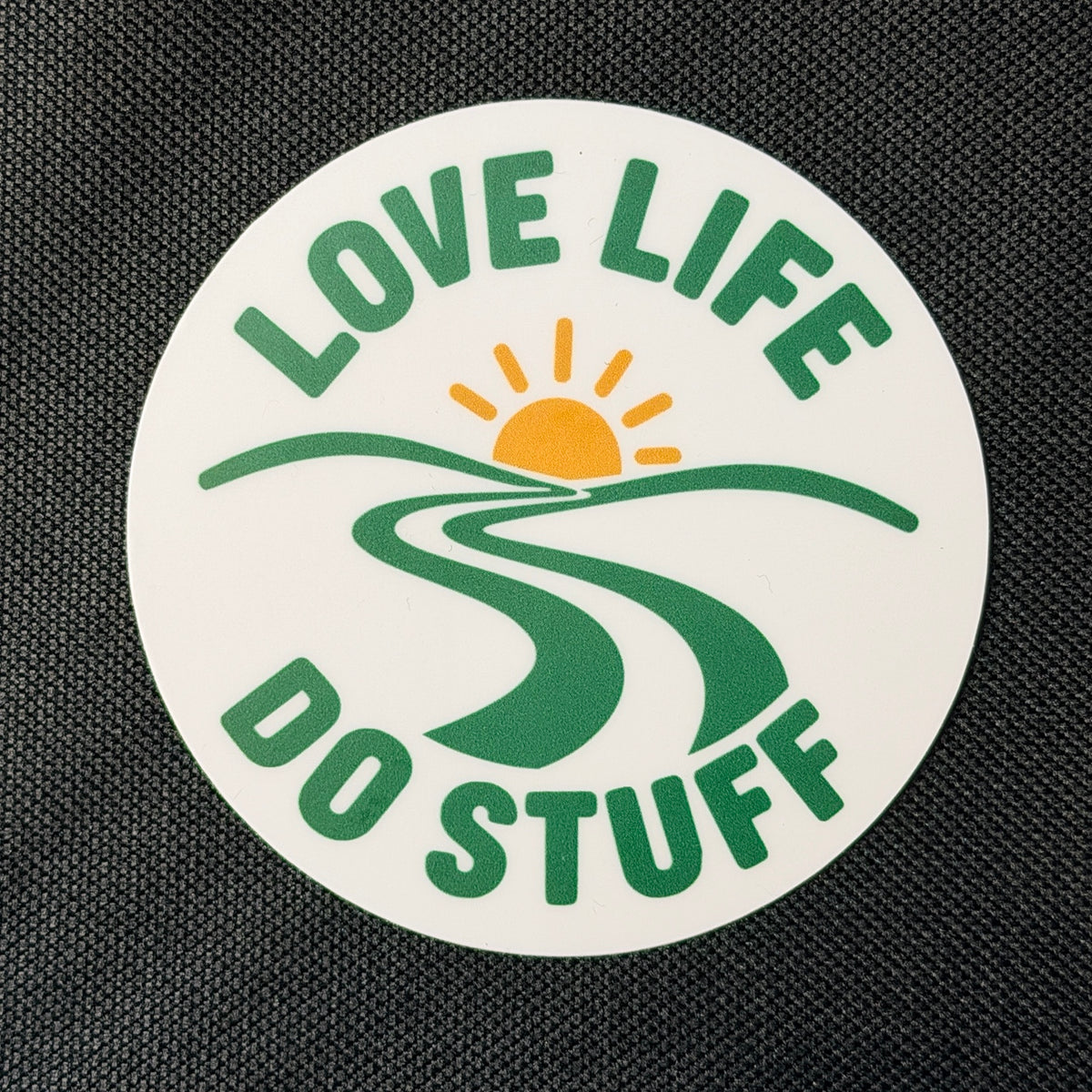 Round Wanderlust Badge vinyl sticker on black fabric, featuring green Love Life Do Stuff lettering, a winding green path, and a golden sun on a white background.